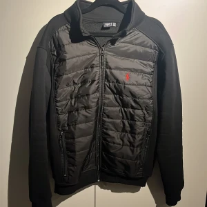 Ralph Lauren Cardigan - Svart pufferjacka/ Cardigan från Polo Ralph Lauren med quiltad framsida och broderad röd logga på bröstet. Jackan har dragkedja framtill, två sidofickor med dragkedja och mjuka ärmar och rygg i bomullsmix. Perfekt för kyliga dagar.
