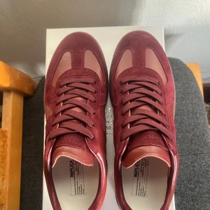 Maison Margiela vinröda sneakers - Maison Margiela sneakers i vinrött med snygga detaljer i mocka och skinn. Klassisk siluett med rund tå, platt sula och snörning. Clean vibe. Perfekta för dig som vill sticka ut med stilrena och exklusiva sneakers.