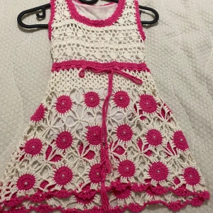 Vit och rosa virkad kortklänning - Superfin kortklänning i virkad stil med vita och rosa färger. Klänningen har ärmlös design, rund hals och ett knytband i midjan. Blommigt mönster med pärldetaljer och volangkant nedtill ger en unik och söt look. Perfekt för sommarens alla tillfällen!