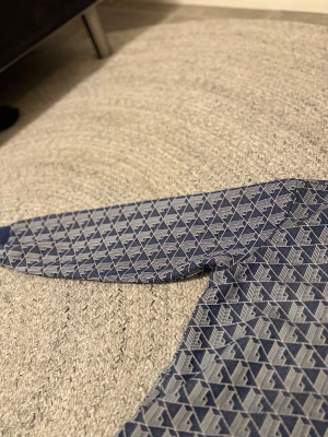 Blå mönstrad zipjacka från Lacoste - Snygg blå zipjacka från Lacoste med vitt geometriskt mönster och klassisk krokodillogga på bröstet. Jackan har ribbade muddar, hög krage och hel dragkedja framtill. Perfekt för en sportig och trendig look.