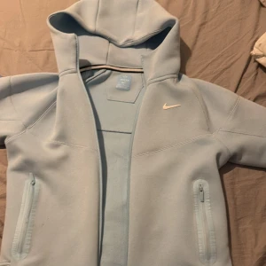 Nike NOCTA ljusblå track suit  - Nike NOCTA tracksuit i ljusblått med dragkedjor på fickorna och vit swoosh-logga på bröstet. Jackan har huva och NOCTA-tryck på ryggen. Tillverkad i mjukt syntetmaterial, perfekt för en chill och sportig look dragkedjan är sönder