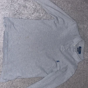Grå långärmad pikétröja Polo Ralph Lauren - Snygg grå långärmad pikétröja från Polo Ralph Lauren i slim fit. Riktigt stilrena tröja som passar perfekt till jeans eller chinos. Tröjan har ett minimalt hål i armhålan men annars riktigt fint skick. Modellen är 177 och väger ~65kg och passar perfekt.
