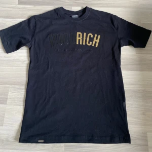 Svart Hoodrich t-shirt med guldtryck - Svart t-shirt från Hoodrich i bomull med stort tryck i svart och guld på bröstet. Klassisk rund hals och korta ärmar. Snygg streetwear-stil med cool branding framtill. Perfekt för dig som gillar trendiga och bekväma plagg.