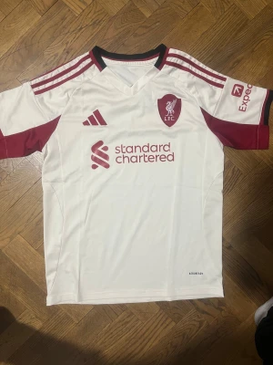 Liverpool FC  - Liverpool FC bortatröja från Adidas i vit med vinröda detaljer och logga. Tröjan har korta ärmar, klassisk V-ringning och tre ränder på axlarna. Tillverkad i lätt och ventilerande polyester med Aeroready-teknologi. Perfekt för dig som älskar fotboll och vill sticka ut.