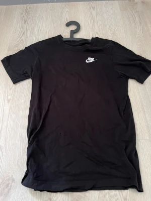 Svart Nike t-shirt i bomull XL (barn) - Svart t-shirt från Nike i storlek XL, med klassisk vit Nike-logga på bröstet. Modellen har rund hals och korta ärmar, tillverkad i mjuk bomull som är skön mot huden. Perfekt för dig som gillar enkel och sportig stil.