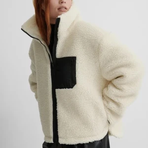 Vit teddyjacka från NA-KD - Säljer en vit teddyjacka från NA-KD (slutsåld) med svart dragkedja och svart bröstficka. Jackan har hög krage, lång ärm och är gjord i mjuk fleece som ger en riktigt mysig vibe. Perfekt för kyliga dagar när du vill vara både varm och snygg.
