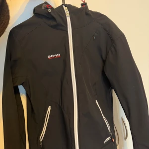 Svart softshelljacka 8848 Altitude - Svart softshelljacka från 8848 Altitude med vit dragkedja och detaljer. Jackan har huva, två fickor med dragkedja och broderad logga på bröstet. Materialet är tekniskt och vindtätt, perfekt för utomhusaktiviteter. Snygg och sportig look med smidig passform.