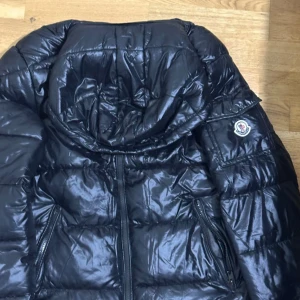 Svart Moncler pufferjacka M - Säljer en svart pufferjacka från Moncler i storlek M. Jackan har glansig finish, dragkedja framtill, två fickor med dragkedja och klassisk Moncler-logga på ärmen. Perfekt för kalla dagar med sin vadderade design och hög krage.
