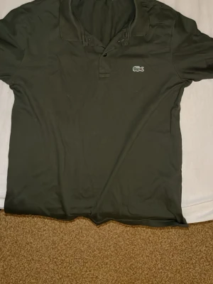 Mörkgrön pikétröja från Lacoste - Säljer en mörkgrön pikétröja från Lacoste med klassisk krage och två knappar framtill. Tröjan har den ikoniska krokodilloggan broderad på bröstet och är tillverkad i mjuk bomull. Perfekt till en avslappnad men ändå stilren look.