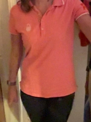 Rosa pikétröja från Sir of Sweden - Rosa pikétröja med vit och orange broderade detaljer på ärmarna, bland annat siffran 77 och texten SIR. Tröjan har klassisk krage, korta ärmar med randig mudd och knappar framtill. Materialet är mjuk bomull och tröjan har en broderad logga på bröstet.