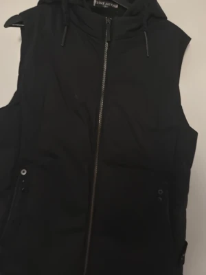 Svart dunväst från Stone Island - Snygg svart dunväst från Stone Island med huva och dragkedja framtill. Västen har två fickor med knappstängning och klassisk logga på sidan. Perfekt för lager-på-lager och streetwear-vibe.