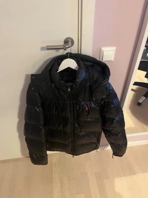 Svart glansig pufferjacka från Polo Ralph Lauren - Säljer en svart pufferjacka från Polo Ralph Lauren i storlek M. Jackan har huva, dragkedja framtill och Polo-logga i rött på bröstet. Den är vadderad och har en glansig finish som ger en cool look. Perfekt för kalla dagar när du vill hålla stilen. Den har en liten skada på ena ärmen. Den är fixad men syns lite om man kollar ordentligt 