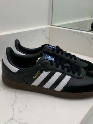 Adidas Samba svart/vit sneakers - Klassiska Adidas Samba sneakers i svart skinn med vita detaljer och de ikoniska tre ränderna på sidorna. Skorna har brun gummisula, svart snörning och blå Adidas-logga på plösen. Perfekt för dig som gillar retrostil och streetwear.( Äkta morsan jobbar där)