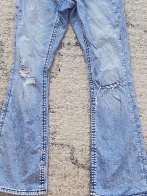 Ljusblå bootcut jeans med slitningar - Säljer ett par ljusblå jeans med bootcut-modell och slitna detaljer på båda benen. Jeansen har klassisk femficksdesign och är tillverkade i mjukt denimtyg. Perfekta för dig som gillar en avslappnad och trendig look.