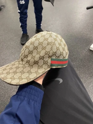 Gucci keps - Säljer en beige keps från Gucci med klassiskt GG-monogram över hela kepsen och en grön-röd rand på sidan. Kepsen är tillverkad i canvasliknande material och har justerbar passform. Perfekt för dig som vill ha en ikonisk och stilren accessoar.