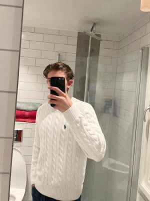 Vit kabelstickad tröja från Ralph Lauren - Säljer en klassisk vit kabelstickad tröja från Ralph Lauren med rund hals och broderad logga på bröstet. Tröjan har långa ärmar och ribbade muddar, perfekt för en clean och stilren look.