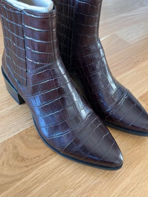 Bruna boots i crocopräglat skinn Vagabond - Säljer ett par mörkbruna boots från Vagabond i crocopräglat skinn. Modellen har spetsig tå, låg klack och en stilren siluett. Perfekta för dig som vill ha ett par boots med lite edge och snygg finish. Inköpspris 1099 :- från vagabond. 