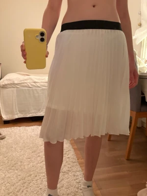 Vit plisserad kjol från Voyelles - Säljer en vit plisserad kjol från Voyelles i storlek S. Kjolen har ett svart elastiskt midjeband och är gjord i 100% polyester. Den har en luftig känsla och slutar strax ovanför knäna, perfekt för en fräsch och stilren look. Hör av dig om du undrar mått eller annat så löser jag det. Använd någon enstaka gång. 