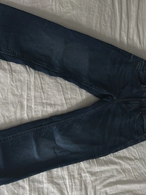 Blå jeans från Lindex, rak modell - Säljer ett par klassiska blå jeans från Lindex i rak passform. Jeansen har fem fickor, bälteshällor och knapp med dragkedja. Materialet är tåligt denim i bomull och färgen är mörkblå med lätt tvättade detaljer. Perfekta till vardags och enkla att matcha.