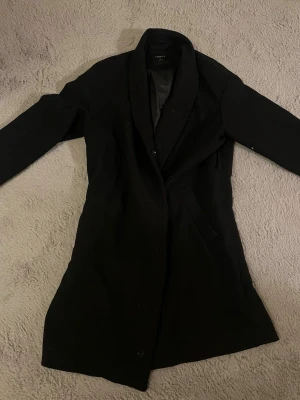 Svart kappa från Lindex, stl S - Säljer en svart, klassisk kappa från Lindex i storlek S. Jackan har enkel knäppning framtill, två fickor och en rak, stilren passform. Materialet är mestadels polyester med inslag av viskos och elastan för extra komfort. Perfekt för kyligare dagar. Det finns lite katthår på den och knappen har bytts ut,  vilket syns på bild 2. Den är stor i storleken och passar M och säkert L. Hade varit snyggare med ett bälte i midjan så rekommenderar att köpa det om man inte redan har ett 🫶🏻 