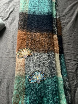 Rutig ullväst med broderade blommor - Unik rutig ullväst i brunt, beige, svart och turkos med stora broderade lotusblommor i olika färger. Västen har en mjuk, fluffig känsla och ett oversized uttryck. Perfekt för dig som vill sticka ut med coola detaljer och färgkombinationer.