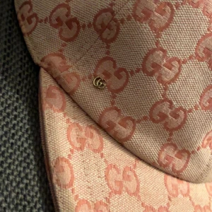  rosa Gucci keps - Snygg keps från Gucci i beige och rosa med klassiskt GG-mönster över hela kepsen. Justerbar rem bak i rosa skinn och liten guldfärgad GG-detalj på sidan. Materialet är bomull och skinn, perfekt för dig som vill sticka ut med lyxig streetstyle.