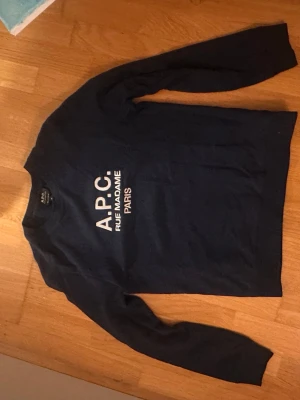 Mörkblå sweatshirt från A.P.C. - Säljer en mörkblå sweatshirt från A.P.C. med vit text 'A.P.C. RUE MADAME PARIS' på bröstet. Tröjan har rund halsringning och långa ärmar. Tillverkad i mjuk bomull, perfekt för en clean och stilren look.