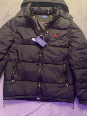 Svart pufferjacka från Polo Ralph Lauren - Säljer en svart pufferjacka från Polo Ralph Lauren med huva och flera dragkedjeförsedda fickor. Jackan har det klassiska röda Polo-logotypen broderad på bröstet och är tillverkad i ett glansigt syntetmaterial. Perfekt för kalla dagar och riktigt snygg streetstil. Den är storlek L men den passar storlek M.