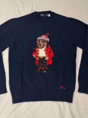 Mörkblå stickad tröja Polo Bear RL - Mörkblå stickad tröja från Polo Ralph Lauren med ikoniska Polo Bear-motivet framtill. Björnen har röd jacka, mönstrad mössa och rutiga byxor. RL-broderi nertill. Klassisk rund hals och ribbade muddar. Perfekt för vintern och julen.