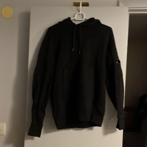 Svart hoodie från C.P. Company - Svart hoodie från C.P. Company med klassisk huva och snörning. Tröjan har lång ärm och ikonisk linsdetalj på vänster ärm. Tillverkad i mjuk bomull för en skön känsla. Storlek S men passar Xs bättre.