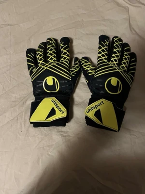 Svarta och gula målvaktshandskar Uhlsport - Säljer ett par svarta och gula målvaktshandskar från Uhlsport med coolt grafiskt mönster och breda kardborreband vid handleden. Handflatan är vit för extra grepp och ovansidan har snygga gula detaljer. Perfekta för dig som älskar fotboll och vill sticka ut på planen.