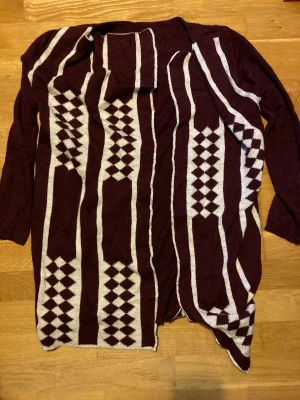 Vinröd och vit kofta med mönster - Säljer en vinröd och vit kofta från MODA S.T. med grafiskt mönster i vitt. Koftan är långärmad och har en öppen front med asymmetrisk nederkant. Materialet är en mix av akryl, alpacka och ull, vilket gör den både mjuk och varm.