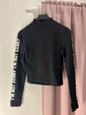 Svart polotopp med text - Säljer en svart polotopp från H&M med långa ärmar. På båda ärmarna står det 'I'M NOT SORRY' i vit text. Toppen är i mjuk bomull och har en tight passform som sitter snyggt. Perfekt för dig som gillar statement-plagg!