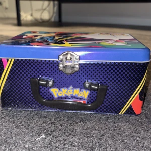Pokémon plåtlåda med kort - Säljer en färgglad Pokémon plåtlåda med motiv av Lucario och Gardevoir på locket. Lådan innehåller flera Pokémonkort, inklusive två oöppnade paket med Basic Energy-kort. Perfekt för samlare eller dig som vill boosta din kortsamling.
