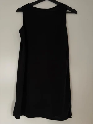 Svart linne från Gatsby - Enkel och stilren svart linne/klänning från Gatsby i kort modell. Linnet är med rund halsringning och har en liten slits i nederkanten för extra detalj. Perfekt att styla med accessoarer för en personlig look. Oanvänt 