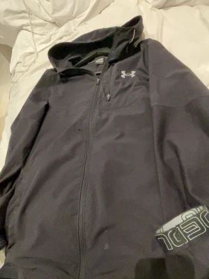 Svart vindjacka från Under Armour - Säljer en svart vindjacka från Under Armour med huva och dragkedja framtill. Jackan har en bröstficka med dragkedja och logga på bröstet. Perfekt för dig som vill ha en sportig och stilren look. Pyre litet hål under bröstet som knappt syns