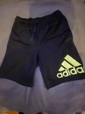 Svarta Adidas Primegreen shorts - Säljer ett par svarta Adidas shorts i Primegreen-material med elastisk midja och snörning. De har en stor gul Adidas-logga på ena benet och är perfekta för träning eller sport. Lätta och bekväma med loose passform.