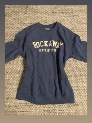Mörkblå sweatshirt - Mörkblå sweatshirt med tryck 'ROCKAWAY BEACH NY 1999' i vitt på bröstet. Klassisk rund halsringning och långa ärmar. Perfekt för dig som gillar college-vibe och avslappnad stil.