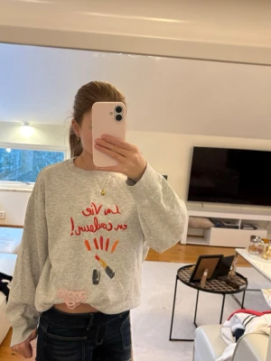 Grå sweatshirt med tryck och läppstift - Säljer en grå sweatshirt med färgglatt tryck framtill som föreställer läppstift och text i rött. Tröjan har en loose passform och långa ärmar, perfekt för en chill och avslappnad stil. Mjuk och skön bomullskänsla.