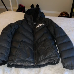 Svart pufferjacka från Nike  - Säljer en svart pufferjacka från Nike i storlek 137/145 för 10-12 år. Jackan har huva, dragkedja framtill och två sidofickor. Stor Nike-logga på bröstet och mönstrat innerfoder. Perfekt för kyliga dagar!Välanvönd men mycket fin. 