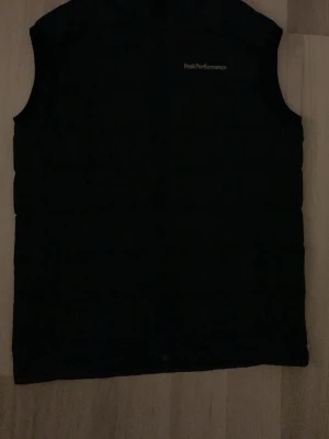 Svart dunväst Peak Performance S - Säljer en svart dunväst från Peak Performance i storlek S. Västen har en stilren design med diskret logga på bröstet och dragkedja framtill. Perfekt att slänga över en hoodie eller tröja när det är kyligt ute.