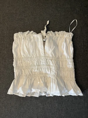 Vit smockad linnetopp med volang - Supersöt vit linnetopp från H&M med smockad resår över hela toppen och volangkant nedtill. Toppen har tunna axelband och en liten knytning framtill. Perfekt för sommardagar och enkel att styla med jeans eller kjol.