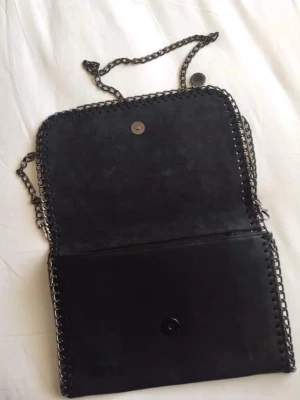 Svart axelväska med kedja - Säljer en svart axelväska i mocka-look med grov silverfärgad kedja som axelrem. Väskan har ett lock med magnetknäppning och dekorativ kedjedetalj runt kanterna. Perfekt storlek för det viktigaste och ger en cool vibe till din outfit.