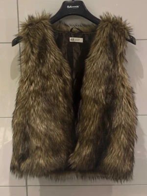 Brun pälsväst från H&M - Säljer en fluffig och snygg brun pälsväst från H&M. Västen har en naturlig look med långa, mjuka fuskpälsstrån och är helfodrad på insidan. Perfekt att slänga över en hoodie eller tröja för extra stilpoäng.