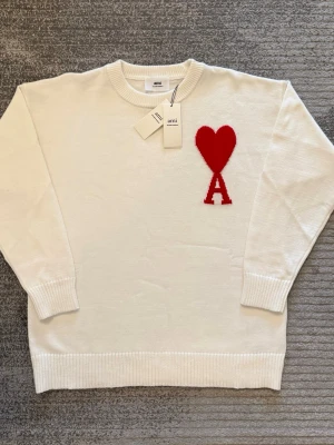 Vit stickad tröja från Ami - Säljer en vit stickad tröja från Ami Alexandre Mattiussi med röd hjärt- och A-detalj på bröstet. Tröjan är gjord av ekologisk bomull, har rund hals och ribbade muddar vid ärmslut och nederkant. Perfekt för dig som gillar stilrena plagg med en twist.