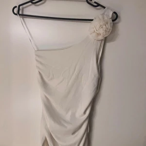 Vit one shoulder topp från NewYorker - Supersnygg vit topp från NewYorker med one shoulder-design och en stor tygblomma på axeln. Toppen har smal passform och är tillverkad i ett mjukt material som faller snyggt. Perfekt för dig som vill sticka ut med en unik detalj på axeln. Aldrig använd!