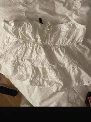 Vit volang topp från Divided - Supersöt vit topp från Divided med tunna axelband och volangdetaljer. Toppen har knyt framtill och är gjord i ett lätt bomullstyg som känns luftigt. Perfekt för varma dagar och enkel att styla med jeans eller kjol.