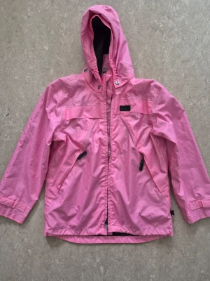 Vindjacka - Ljusrosa vindjacka med huva från Mywear Active. Jackan har dragkedja framtill, två snedställda fickor med dragkedja och justerbara ärmslut. Tillverkad i lätt och tålig polyester, perfekt för blåsiga dagar. Några fläckar.