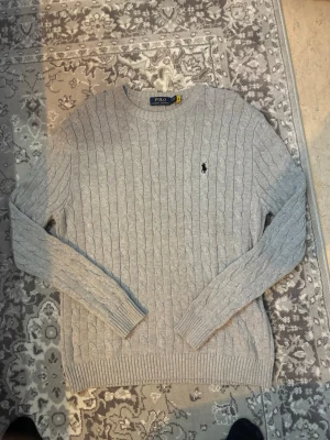 Grå kabelstickad tröja Polo Ralph Lauren - Kabelstickad grå tröja från Polo Ralph Lauren med klassisk rund halsringning och broderad logga på bröstet. Långärmad modell i mjuk bomull som passar perfekt till lager på lager under höst och vinter. Snygg ribbad kant vid ärmslut och nederkant.