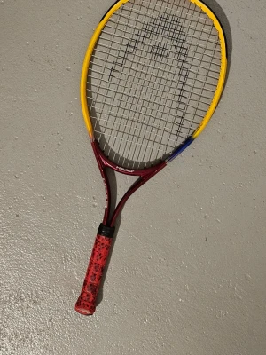 Tennisracket Head Radical Pirate 62. - Modell: Radical Pirate 62, ofta associerad med Andre Agassi-serien.  Design: Racketet har en distinkt design med en färggradient från rött till gult på ramen och ett rött grepp.  Längd: Längden är cirka 62 cm (24,5 tum), 
